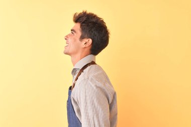 Önlük takmış genç yakışıklı bir işçi, yalıtılmış sarı arka planda duruyor. Yan tarafa bakıyor, rahat bir poz veriyor, kendine güvenen bir gülümsemesi var..