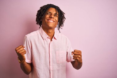Zarif gömlekli Afro-Amerikan bir adam pembe arka planda dikiliyor çok mutlu ve heyecanlı bir şekilde kollarını kaldırıp, gülümseyerek ve başarı çığlıkları atarak kazanan hareketi yapıyor. Kutlama konsepti.