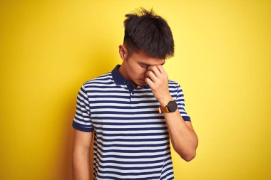 İzole edilmiş sarı arka planda duran çizgili polo giyen Asyalı genç adam yorgun burnu ve gözleri ovuşturarak yorgunluk ve baş ağrısı hissediyor. Stres ve hüsran kavramı.
