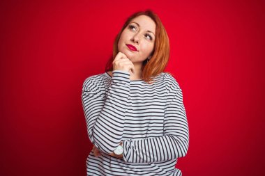 Kızıl saçlı genç bir kadın donanma gömleği giyiyor. Kırmızı izole edilmiş arka plan üzerinde duruyor. Elleri çenesinde, soru sormayı, düşünceli ifadeyi düşünüyor. Düşünceli bir yüzle gülümsüyor. Şüphe kavramı.