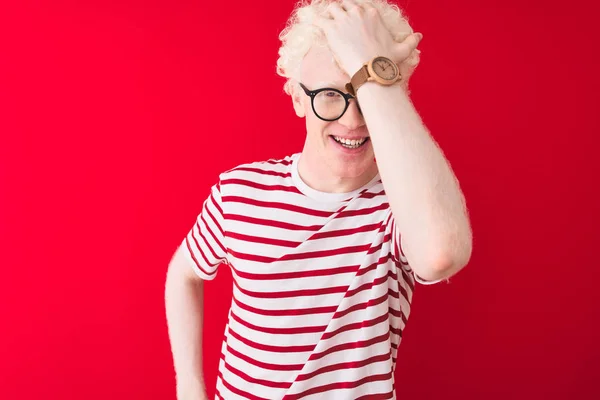 Young albino blond man wearing striped t-shirt and glasses over isolated red background surprised with hand on head for mistake, remember error. Forgot, bad memory concept.