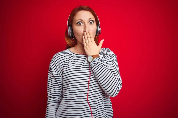 年轻的红头发女人用耳机在红色孤立的背景音乐上 用手捂住嘴听音乐 为自己的错误 恐惧的表情 沉默中的恐惧 神秘的概念而感到羞愧 — 图库照片