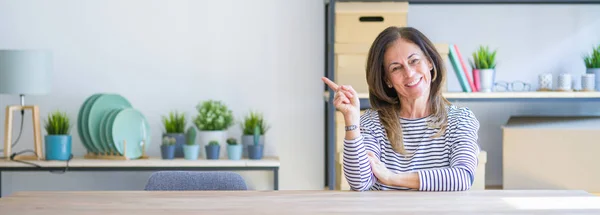 Wide angle photo of middle age senior woman sitting at the table at home with a big smile on face, pointing with hand and finger to the side looking at the camera.