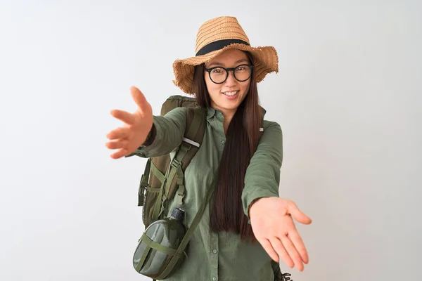 Chinese hiker woman wearing canteen hat glasses backpack over isolated white background looking at the camera smiling with open arms for hug. Cheerful expression embracing happiness.