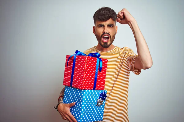 Young man with tattoo holding birthday gifts standing over isolated white background annoyed and frustrated shouting with anger, crazy and yelling with raised hand, anger concept