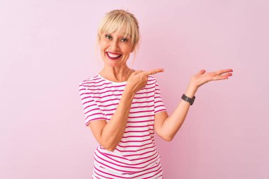 İzole edilmiş pembe arka planda duran çizgili tişört giyen orta yaşlı kadın kameraya hayret ediyor ve gülümsüyor..