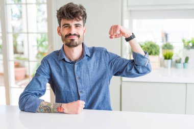 Beyaz masada oturan rahat gömlekli genç adam güçlü biri kol kası gösteriyor, kendinden emin ve gururlu.