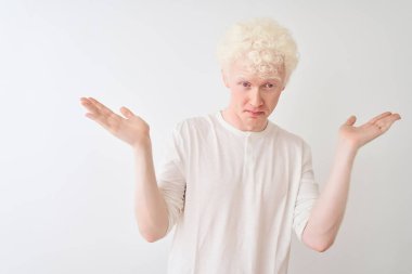 Sıradan tişörtler giyen genç albino bir adam izole edilmiş beyaz arka planda duruyor ve elleri ve kolları havada kafası karışmış bir ifade takınıyor. Şüphe kavramı.