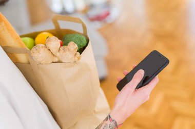 Evde taze bakkaliye dolu bir kağıt çantası tutan genç yakışıklı adam, ürün satın alırken akıllı telefon gösterme ve kullanma