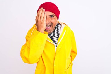 Yağmurluk ve yünlü şapka giyen orta yaşlı bir adam izole edilmiş beyaz arka planda duruyor. Bir gözünü eliyle kapatıyor. Yüzünde kendine güvenen bir gülümseme ve şaşırtıcı bir duygu var..