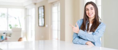 Evdeki beyaz masada oturan güzel genç bir kadının geniş açılı resmi. Yüzünde mutlu ve doğal bir ifadeyle, elleri ve parmaklarıyla gülümseyen bir gülümsemeyle.