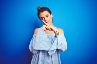 Genç ve güzel bir kadın mavi izole edilmiş arka planda çörek saç stili giyiyor. Reddedilme ifadesi olumsuz işaret veriyor.