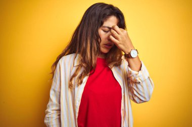 Sarı izole arka plan üzerinde kırmızı tişört ve çizgili tişört giyen genç güzel bir kadın yorgun burnu ve gözleri ovuşturarak yorgunluk ve baş ağrısı hissediyor. Stres ve hüsran kavramı.