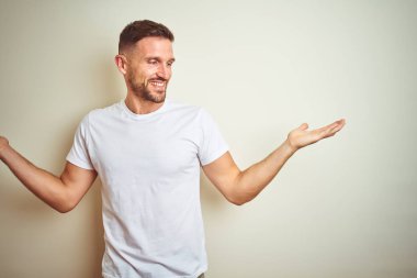 Gülen iki elle gösterilen açık avuç içi, sunma ve karşılaştırma ve denge reklam arka plan üzerinde rahat beyaz t-shirt giyen genç yakışıklı adam izole