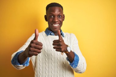 İzole edilmiş sarı arka plan üzerinde kot gömlek ve beyaz kazak giyen Afro-Amerikalı adam el hareketini onaylıyor, gülümsüyor ve başarı için mutlu. Kazanan jest.