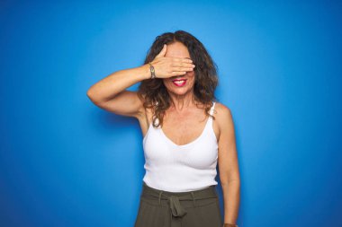 Orta yaşlı, kıvırcık saçlı, mavi izole edilmiş arka planda ayakta duran, gülümseyen ve yüzünde şaşkınlıktan gözleri kaplayan bir gülümsemeyle son sınıf öğrencisi. Kör kavram.