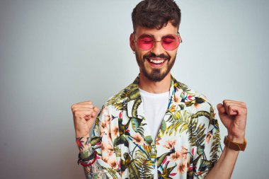 Tatilde dövmeli, güneş gözlüğü takan, izole edilmiş beyaz arka plan üzerinde şaşırmış ve şaşırmış bir şekilde kolları açık bir şekilde başarıyı kutlayan bir adam. Kazanan konsept.