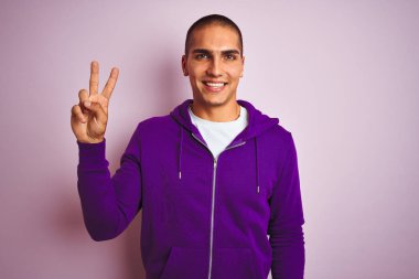 Pembe izole edilmiş arka planda mor eşofman üstü giyen yakışıklı genç adam kameraya gülümseyip zafer işareti yapıyor. İki numara..
