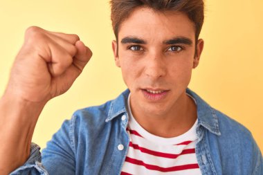 İzole edilmiş sarı arka plan üzerine çizgili t-shirt ve kot gömlek giyen genç yakışıklı adam öfkeyle bağırıyor, çıldırıyor ve öfkeyle bağırıyor.