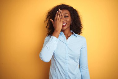 Afro-amerikan iş kadını zarif bir gömlek giyiyor, sarı arka planda, bir gözünü elinle kapatıyor, yüzünde kendine güvenen bir gülümseme ve sürpriz bir duygu var..