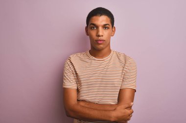 Çizgili tişört giyen genç yakışıklı Arap adam izole edilmiş pembe arka planda duruyor ve yüzünde ciddi bir ifade var. Basit ve doğal bir şekilde kameraya bakıyorsun..