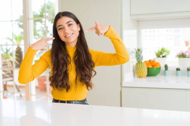 Genç güzel kadın evde kendine güvenen gösterilen ve işaret parmakları diş ve ağız ile gülümseyen beyaz masada. Sağlık kavramı.