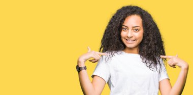 Yüzünde gülümseme ile kendinden emin görünüyorsun, kendini gururlu ve mutlu parmakları ile işaret casual beyaz t-shirt giyen kıvırcık saçlı genç güzel kız.
