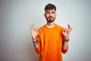 İzole edilmiş beyaz arka plan üzerinde duran turuncu kazak giyen genç adam rahatla ve parmaklarıyla meditasyon hareketi yaparken gözleri kapalı gülümse. Yoga konsepti.