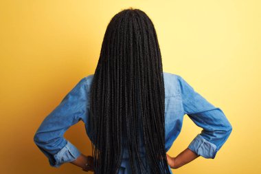 Kot gömlek giyen genç Afrikalı Amerikalı kadın izole edilmiş sarı arka planda dikiliyor geriye bakıyor kollarını vücuduna dikiyor.
