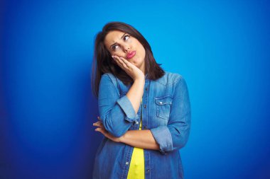 Genç, güzel, esmer bir kadın mavi kot ceket giyiyor. Mavi izole bir arka plan. Kolları çaprazlanmış depresyondan sıkılmış ve yorgun görünüyor..