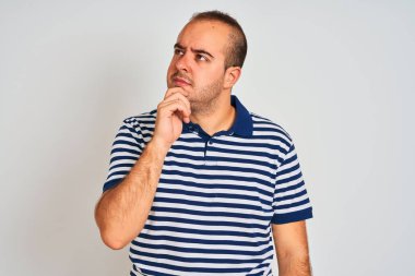 Sıradan çizgili polo giyen genç bir adam izole edilmiş beyaz arka planda duruyor, elleri çenesinde soru sorarken, düşünceli bir ifade takınıyor. Düşünceli bir yüzle gülümsüyor. Şüphe kavramı.