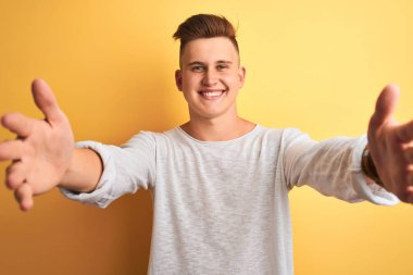 İzole edilmiş sarı arka plan üzerinde duran beyaz tişörtlü genç yakışıklı adam sarılmak için kollarını açmış gülümseyen kameraya bakıyor. Mutluluğu kucaklayan neşeli bir ifade..