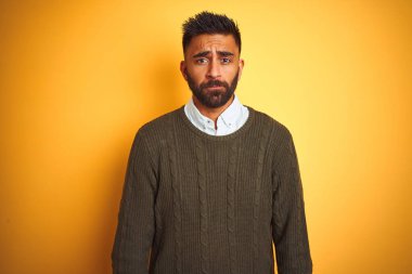 Yeşil kazak ve tişört giyen genç Hintli adam izole edilmiş sarı arka planda duruyor. Depresyonda ve endişe içinde, sinirli ve korkmuş bir halde. Üzücü ifade.