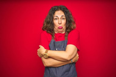 Kırmızı izole arka planda önlük giyen orta yaşlı bir kadın kış soğuğu yüzünden titriyor ve donuyor. Yüzünde üzgün ve şok ifadesi var.