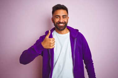 Mor eşofman altı giyen genç Hintli adam pembe arka planda dikiliyor ve elleriyle mutlu bir jest yapıyor. Kameraya bakıp başarıyı gösteren ifade onaylanıyor.