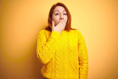 Kışlık süveter giyen güzel kızıl saçlı kadın izole edilmiş sarı arka planda dikiliyor. Elle ağzını kapatmaktan sıkılmış. Huzursuz ve uykulu..