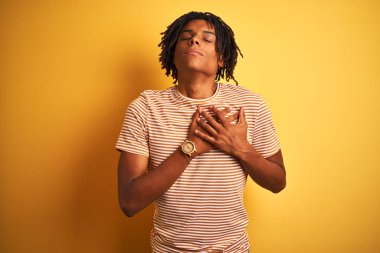 Sarı arka plan üzerinde çizgili t-shirt giyen Afro bir adam elleri göğsünde, gözleri kapalı ve yüzünde minnet dolu bir jest ile duruyor. Sağlık kavramı.