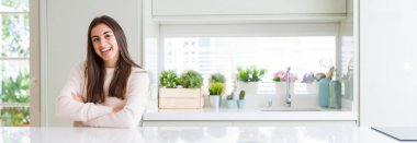 Evde beyaz masada oturan güzel genç kadının geniş açılı resmi kameraya bakarak çapraz kollarıyla gülümseyen mutlu bir yüz. Pozitif kişi.