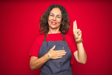 Kırmızı izole edilmiş arka planda önlük giymiş orta yaşlı bir kadın gülümsüyor, elleri göğsünde ve parmakları havada, sadakat yemini ediyor.