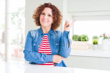 Orta yaşlı, kıvırcık saçlı, kot ceket giyen, yüzünde kocaman bir gülümseme olan, eli kameraya bakan, orta yaşlı bir kadın..