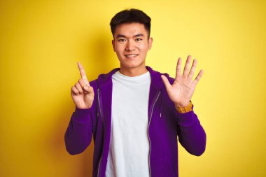 Mor kazak giyen Asyalı genç adam izole edilmiş sarı arka planda duruyor. Altıncı parmaklarını gösterip gülümserken kendinden emin ve mutlu görünüyor..