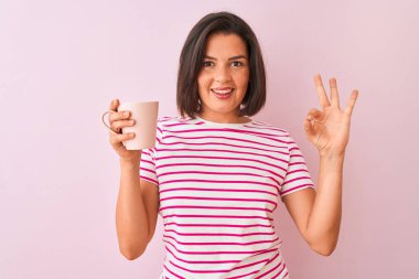 Bir fincan kahve içen genç ve güzel bir kadın izole edilmiş pembe arka planda duruyor. Tamam işareti yapıyor, mükemmel bir sembol.