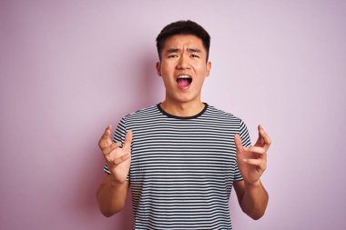 Çizgili tişört giyen Asyalı genç adam pembe arka planda dikiliyor. Deli gibi bağırıyor ve agresif bir ifade ve kollarla bağırıyor. Hayal kırıklığı konsepti.