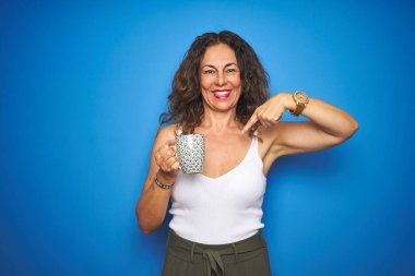 Orta yaşlı bir kadın mavi izole bir arka planda bir fincan kahve içiyor.