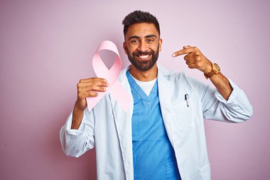 Kanser kurdelesi tutan genç Hintli doktor soyutlanmış pembe arka planda el ve parmak göstererek çok mutlu duruyor.