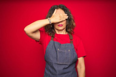 Kırmızı izole edilmiş arka planda önlük giyen orta yaşlı bir kadın. Gözleri elle kapatıyor, ciddi ve üzgün görünüyor. Görme, saklama ve reddetme konsepti