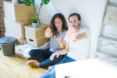 Orta yaşlı bir çift yeni bir eve taşınıyor. Akıllı telefon kullanıyorlar ve ellerinde dur işareti var. Ciddi ve kendinden emin bir ifade var.