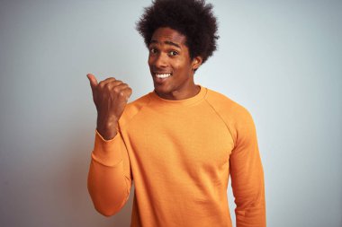Afro saçlı, turuncu süveter giyen, izole edilmiş beyaz arka planda gülümseyen, gülümseyen ve baş parmağıyla yüzünü gösteren genç bir Amerikalı..