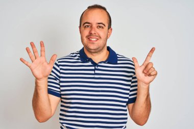Sıradan çizgili polo giyen genç adam izole edilmiş beyaz arka planda duruyor ve yedinci parmaklarıyla işaret ederken kendinden emin ve mutlu bir şekilde gülümsüyor..