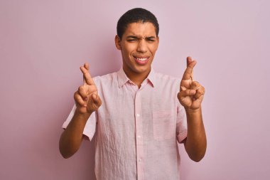 İzole edilmiş pembe arka plan jestlerinin üzerinde dikilen rahat gömlekli genç yakışıklı Arap adam umut ve gözler kapalı gülümseyerek el sallıyor. Şans ve batıl inanç..